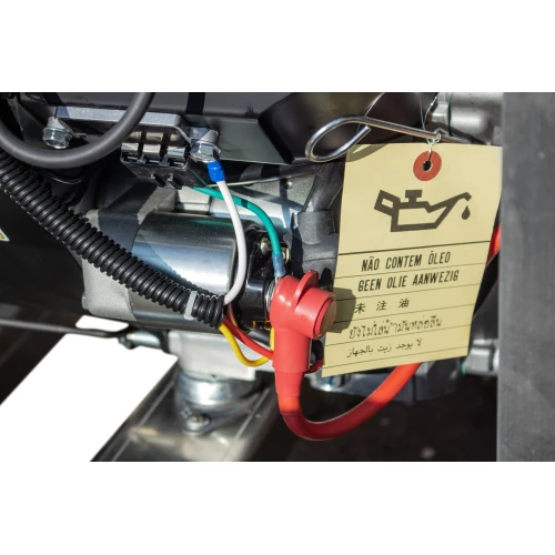 Zbliżenie na część agregatu prądotwórczego z widocznymi przewodami i oznaczeniem ostrzegawczym dotyczącym oleju. Na etykiecie znajdują się instrukcje w kilku językach oraz symbol oleju.