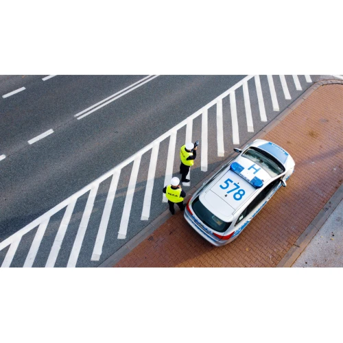 Na zdjęciu widać dwóch policjantów w kamizelkach odblaskowych stojących obok policyjnego samochodu zaparkowanego na poboczu drogi. Jeden z policjantów trzyma alkomat, a drugi obserwuje sytuację na ulicy.