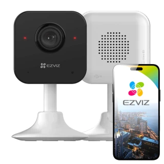Na zdjęciu znajduje się czarna kamera domowa WiFi EZVIZ H1C na białej podstawce, obok której stoi smartfon z wyświetlonym logo EZVIZ i widokiem miasta. W tle widać tylną część kamery z otworami wentylacyjnymi.