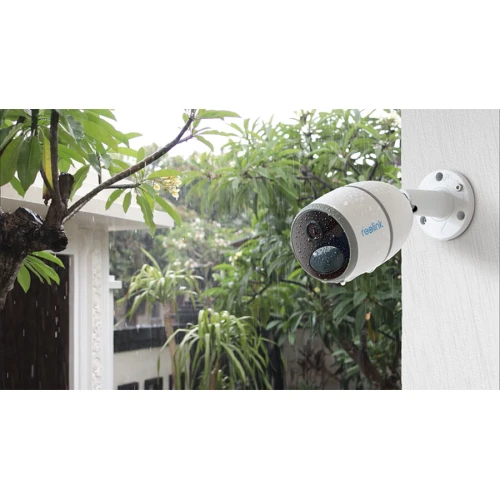 Kamera zewnętrzna IP Reolink jest zamontowana na ścianie budynku, skierowana w stronę ogrodu. Urządzenie ma cylindryczny kształt i jest odporne na warunki atmosferyczne, co widać po kroplach deszczu na jego obudowie.