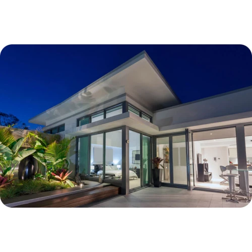 Nowoczesny dom z dużymi przeszkleniami i oświetleniem zewnętrznym, otoczony roślinnością. Wnętrze widoczne przez szklane drzwi prezentuje elegancki, minimalistyczny wystrój.