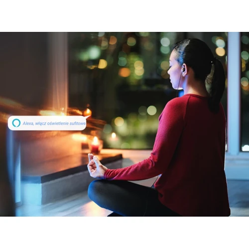 Na zdjęciu znajduje się osoba siedząca w pozycji medytacyjnej w przyciemnionym pomieszczeniu, z zapalonymi świecami w tle. Obok widoczny jest tekst sugerujący użycie asystenta głosowego do włączenia oświetlenia sufitowego.