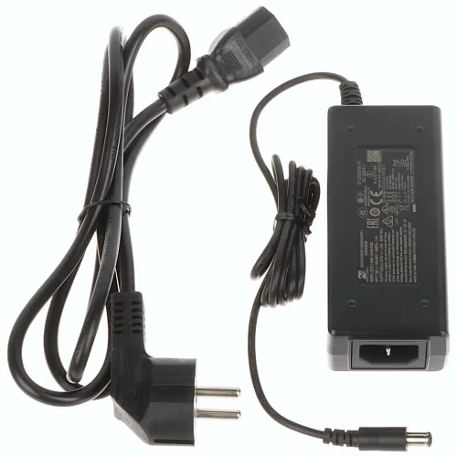 Na zdjęciu znajduje się czarny zasilacz z przewodem zasilającym zakończonym wtyczką oraz przewodem wyjściowym z okrągłą końcówką. Zasilacz ma prostokątny kształt z widocznymi oznaczeniami technicznymi na obudowie.